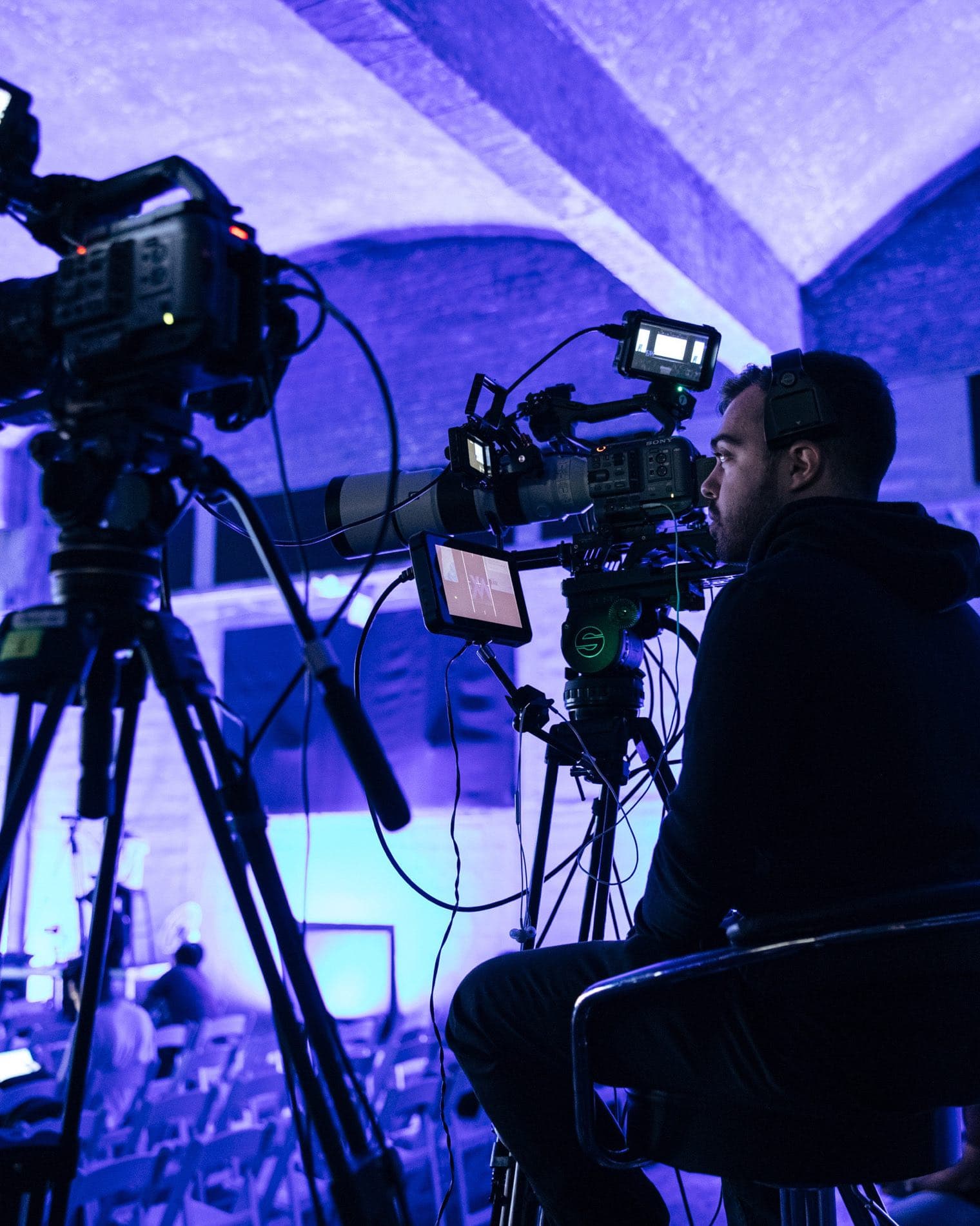 Backlight Media broadcast camera covering the Exness Week stage in a vaulted, purple-lit venue in Cyprus, part of the hybrid livestream linking Cyprus and Kuala Lumpur