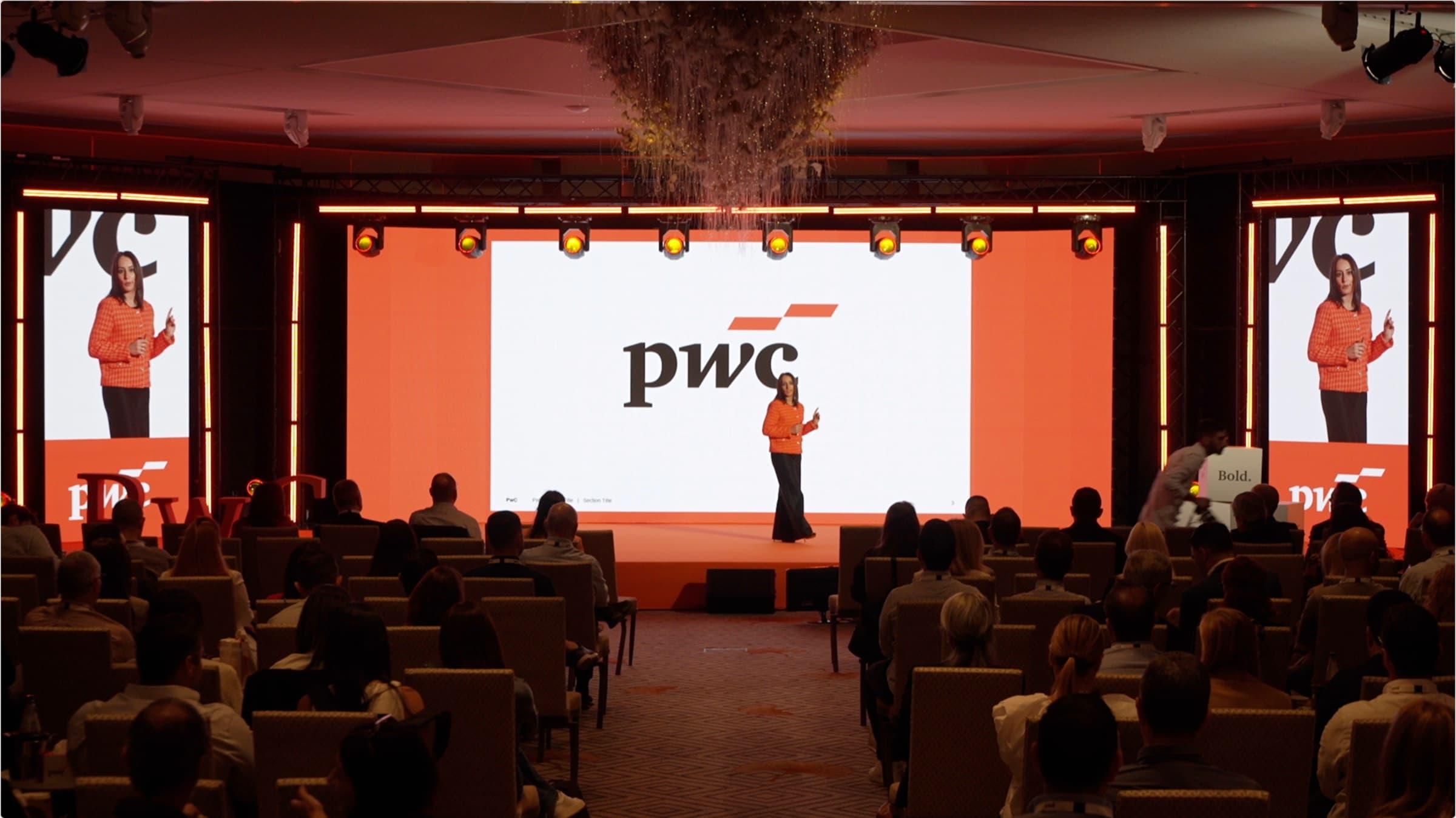 A presenter on the orange-lit main stage at the PwC Annual Conference in Cyprus, the pwc logo on the LED screen and image-magnification side screens, full seated audience, produced by Backlight Media