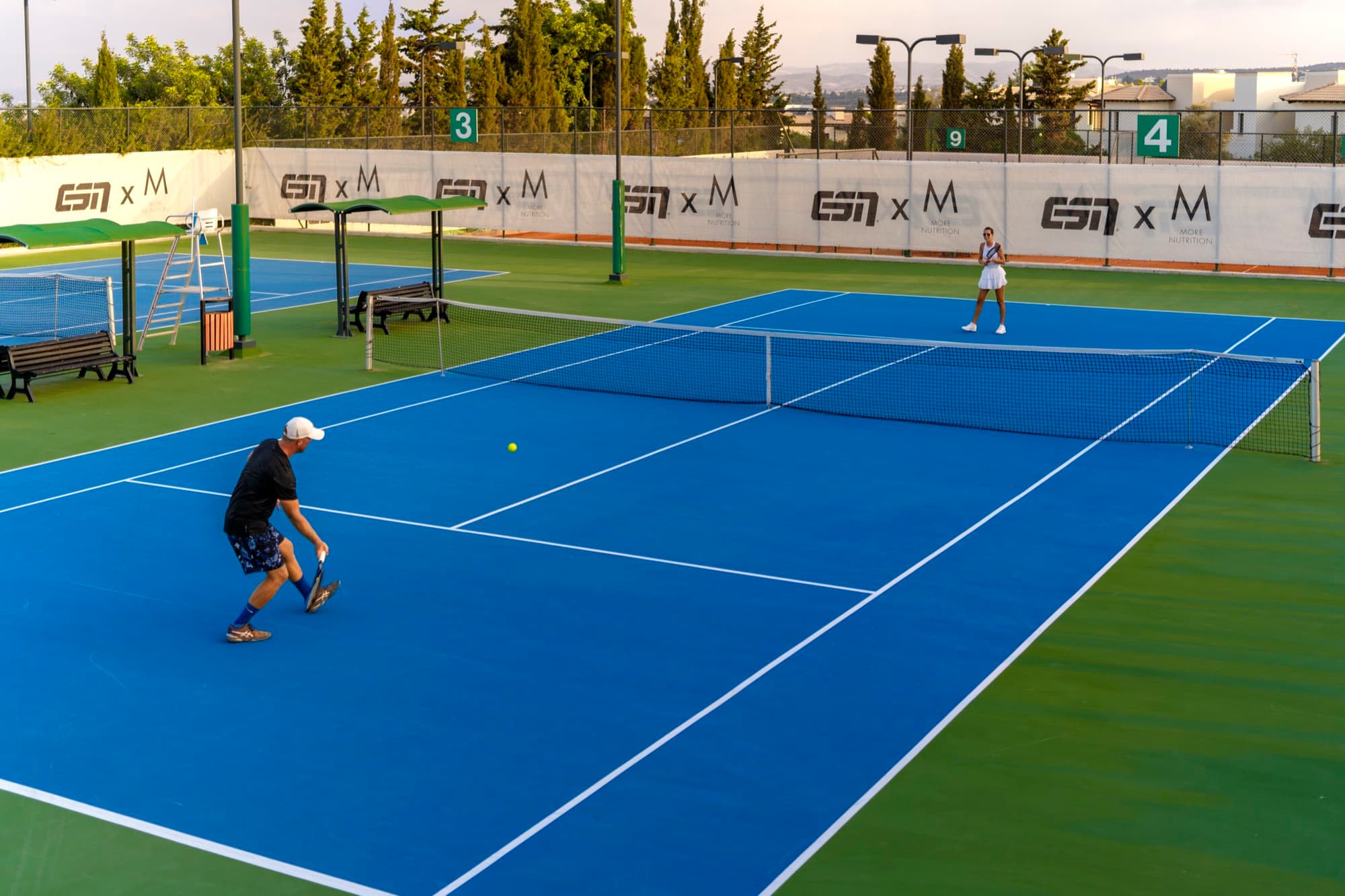 Two players in a rally on the blue tennis hard courts at Aphrodite Hills, captured by Backlight Media