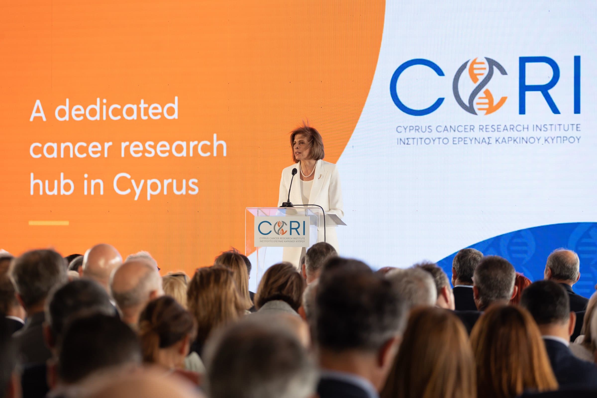 Speaker addressing a full house at the Cyprus Cancer Research Institute (CCRI) grand opening against a wide LED wall reading 'A dedicated cancer research hub in Cyprus', with live AV by Backlight Media