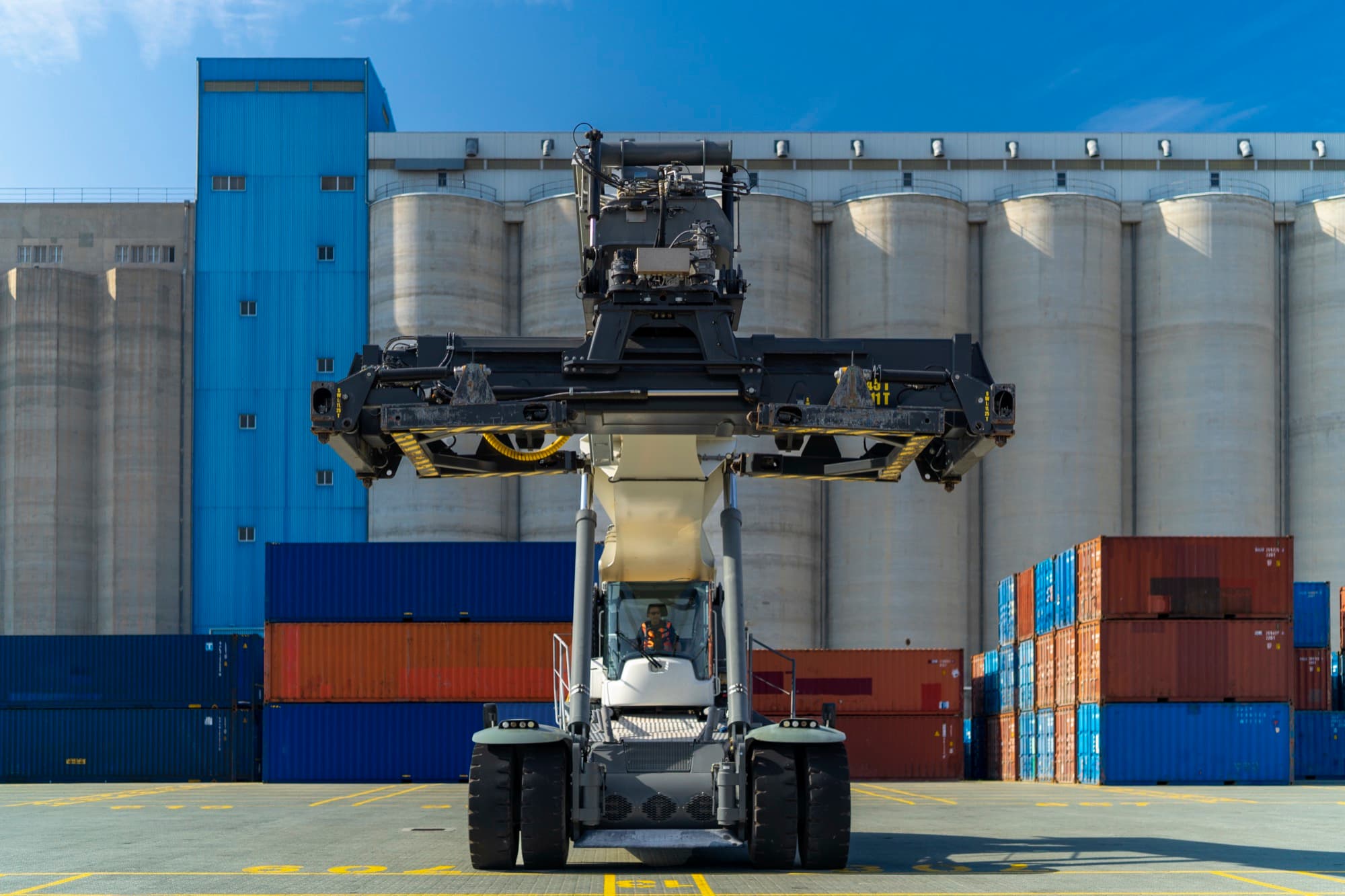 A large black container reach stacker photographed head-on against a grain silo and stacked shipping containers at the DP World Limassol terminal, by Backlight Media