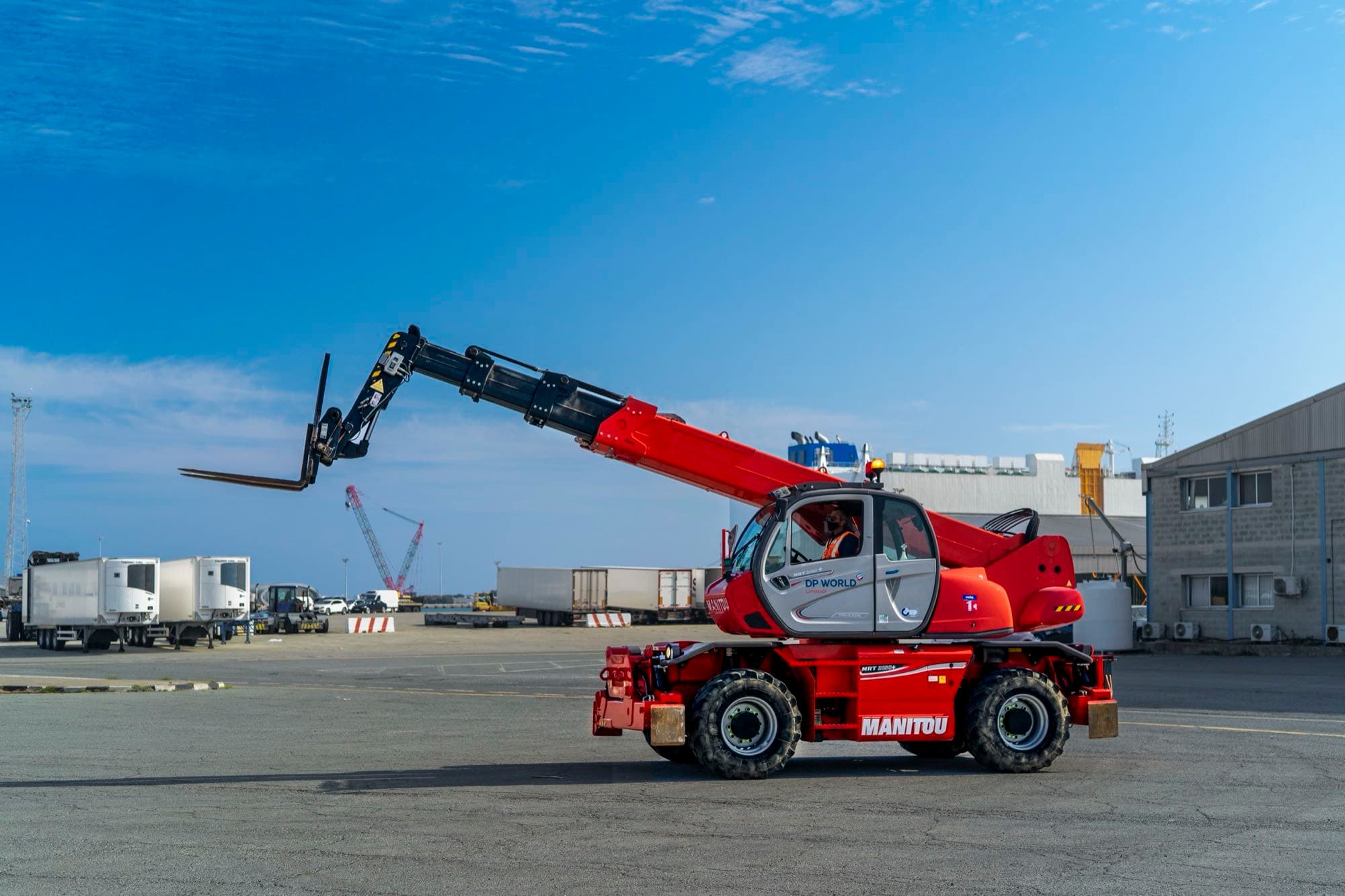 A red DP World Limassol Manitou rotating telehandler with boom extended and an operator in the cab on the port apron, documented by Backlight Media