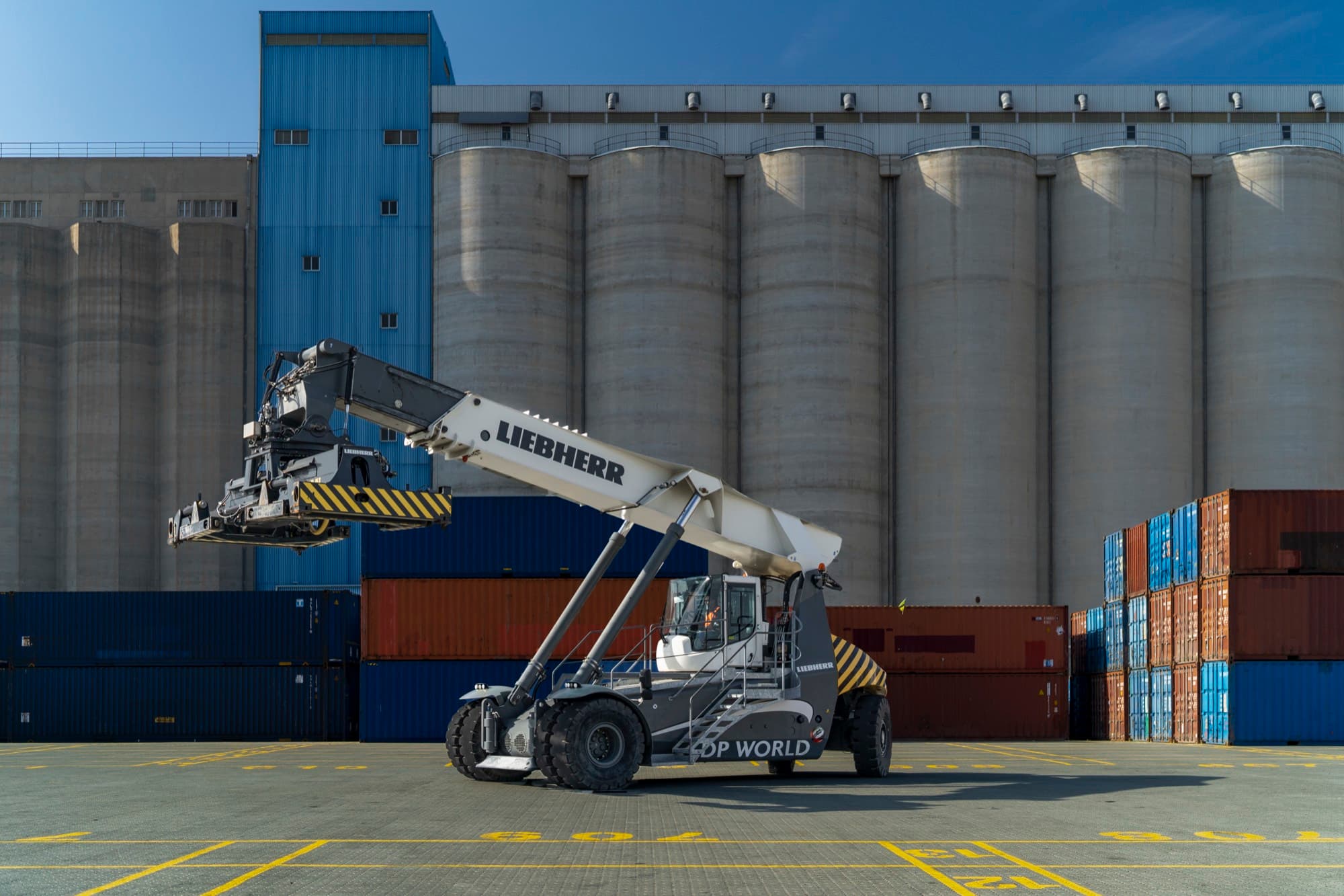 A Liebherr reach stacker in DP World livery with its boom raised against tall concrete grain silos and stacked containers at Limassol port, by Backlight Media