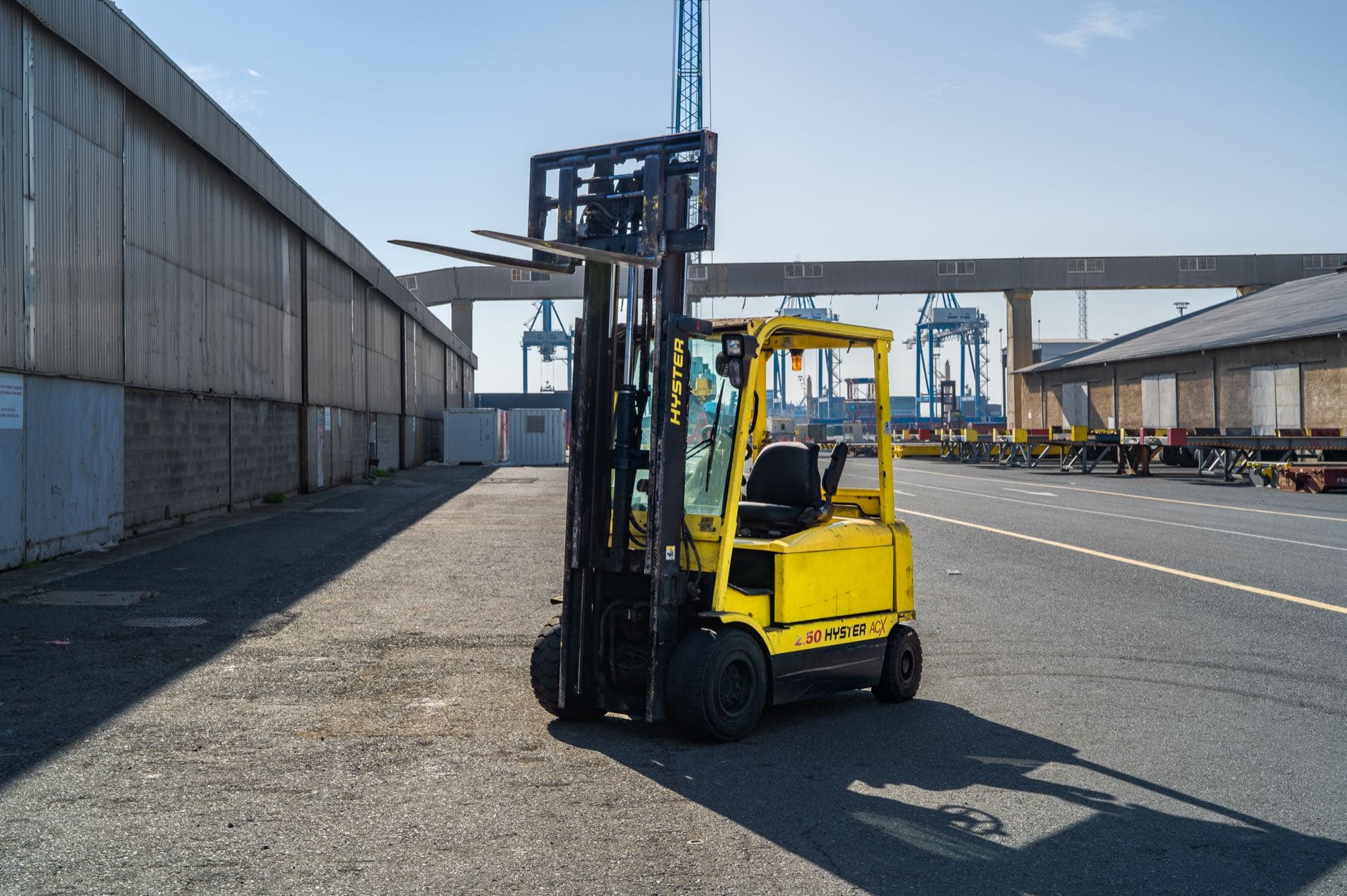 A blue Konecranes reach stacker in DP World livery with telescopic boom raised, a red mobile crane behind on the Limassol port apron, by Backlight Media