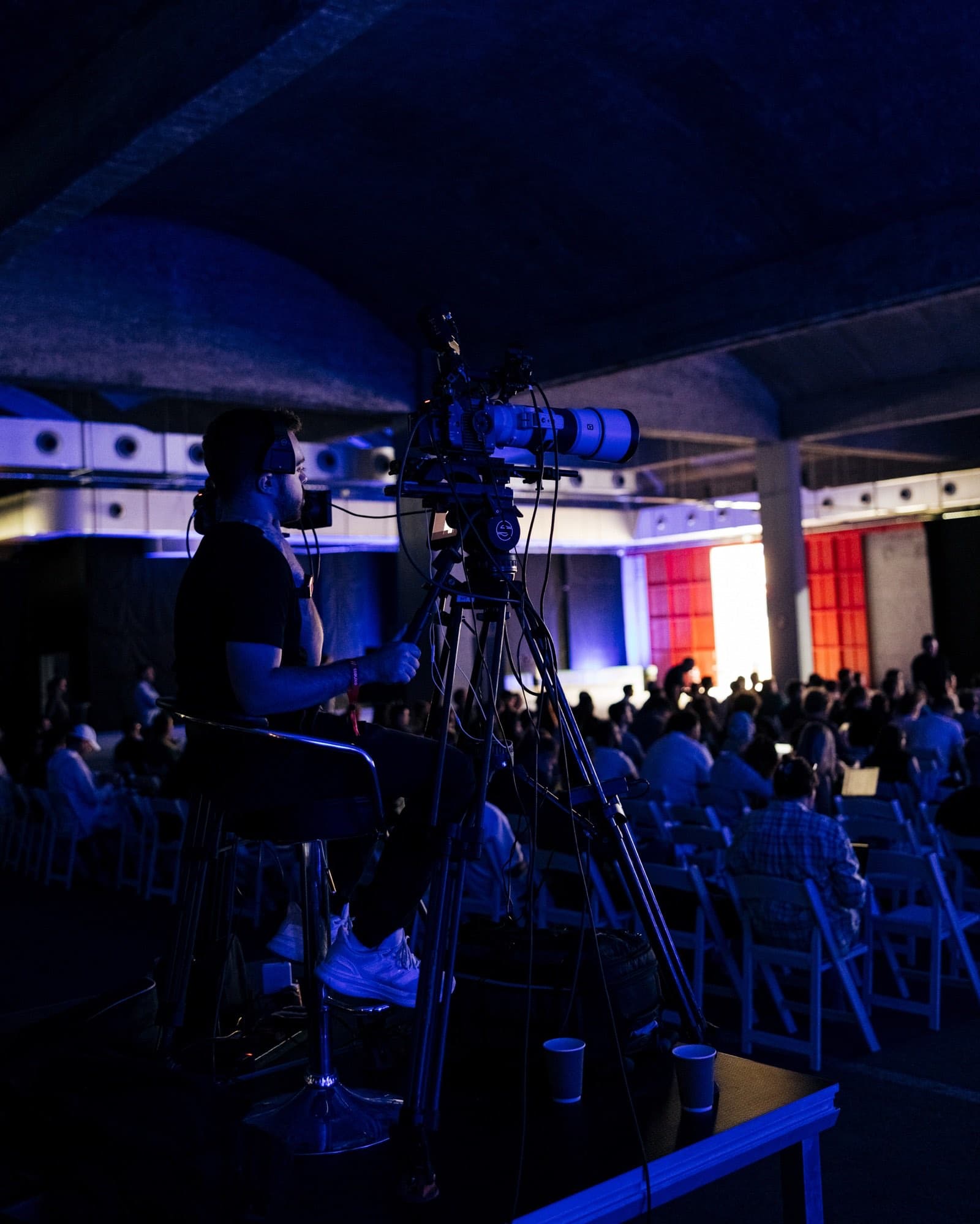 Broadcast cameras on tripods covering the Exness Week stage with the seated audience beyond, in a vaulted, purple-lit venue, filmed by Backlight Media