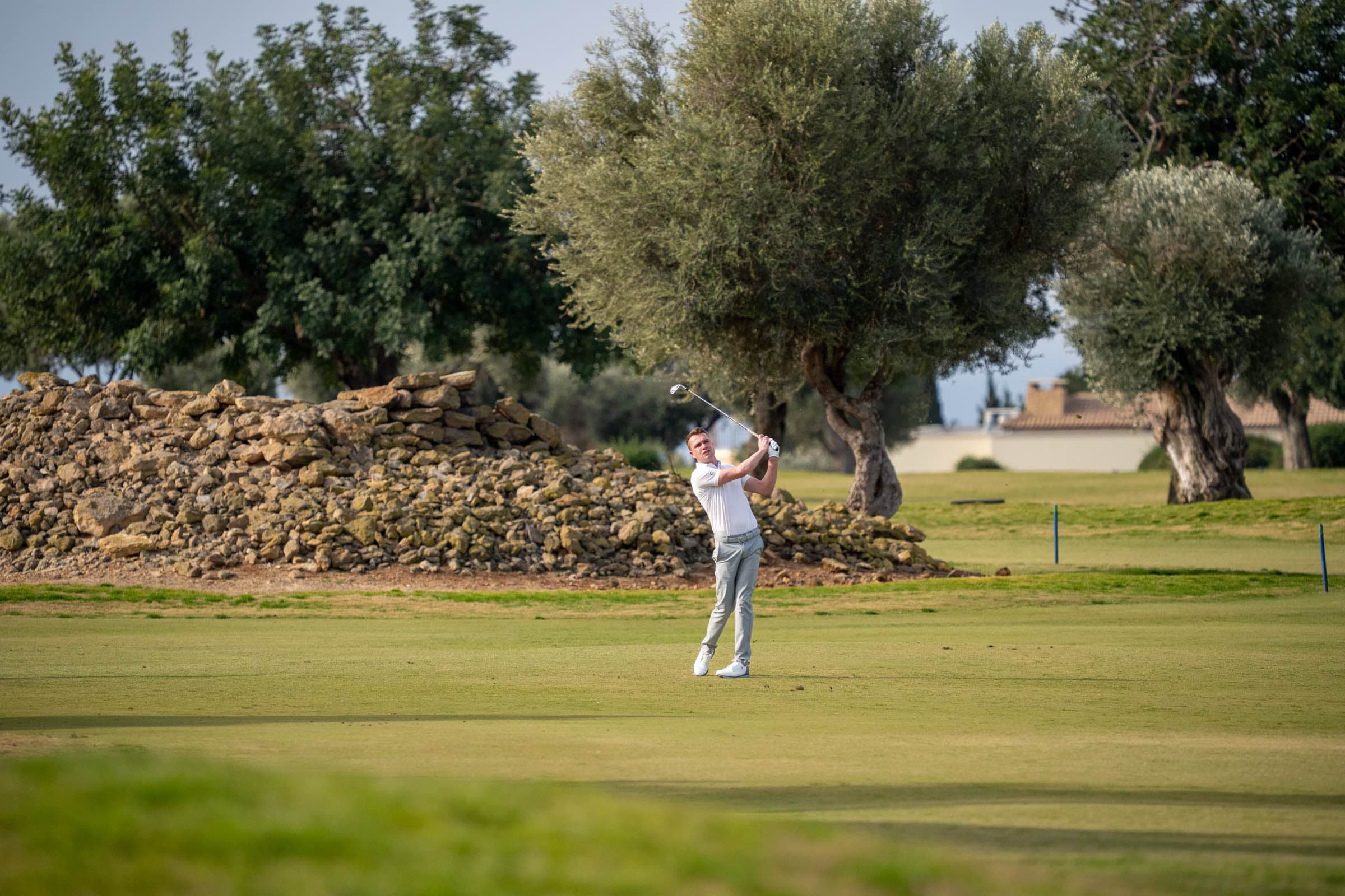 A golfer at full follow-through on the fairway with dry-stone wall and olive trees at the PGA National Cyprus play-offs, Aphrodite Hills, by Backlight Media