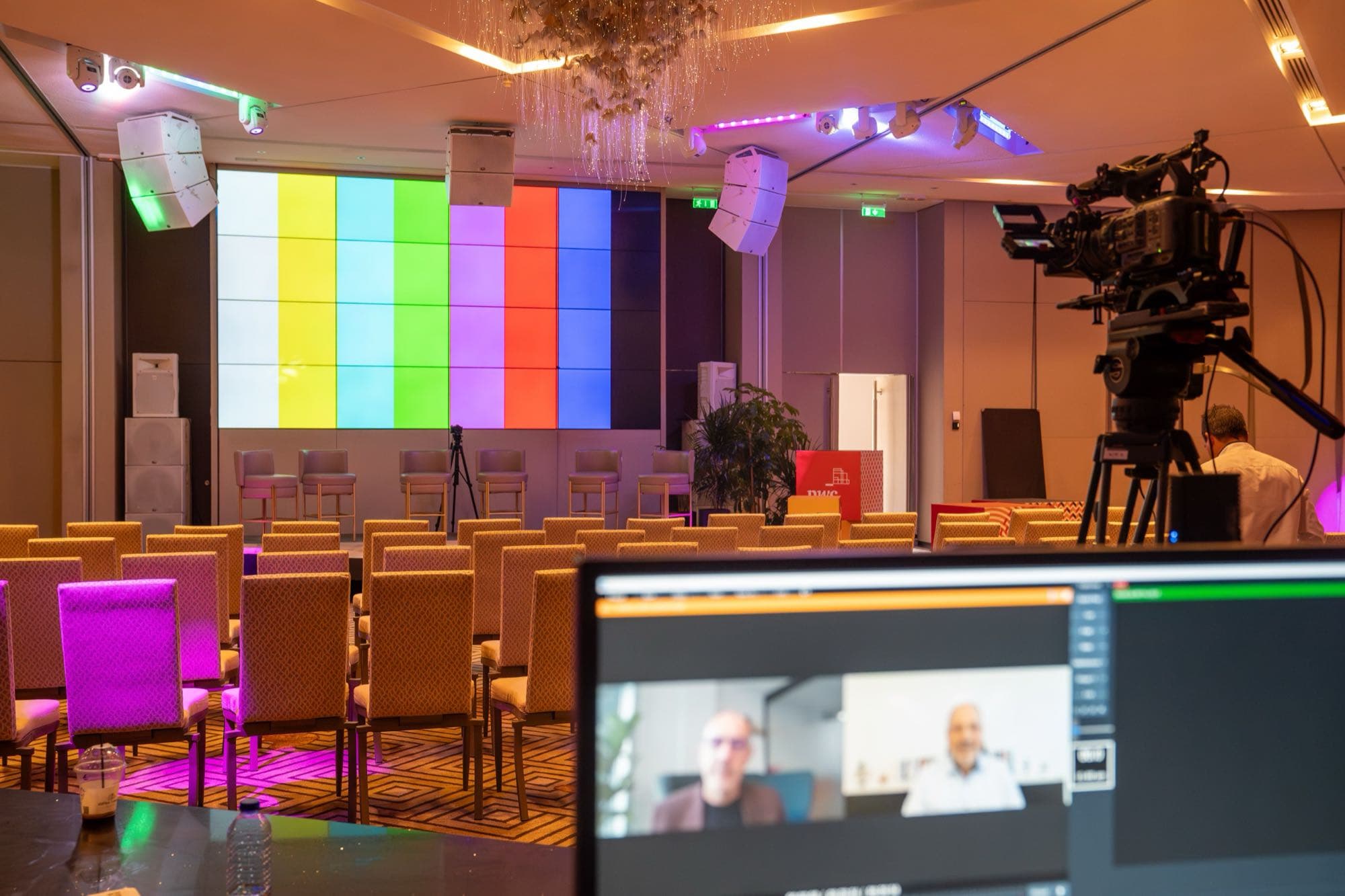 The PwC conference hall set for a hybrid event: LED stage wall, line-array speakers, a broadcast camera and a vision-mixer monitor showing two remote speakers on a video call, by Backlight Media